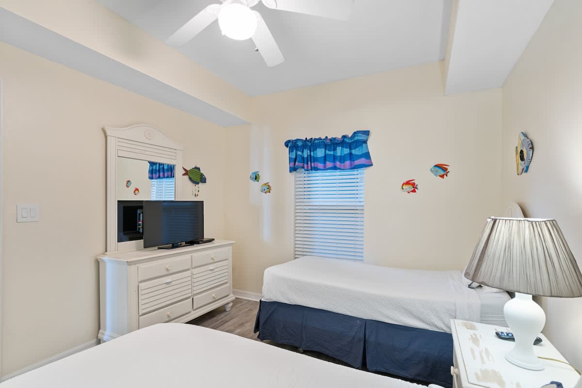 View of the twin bedroom showing a white dresser with a TV and mirror, light wood-look flooring, and a window with blue blinds, continuing the playful fish decor.