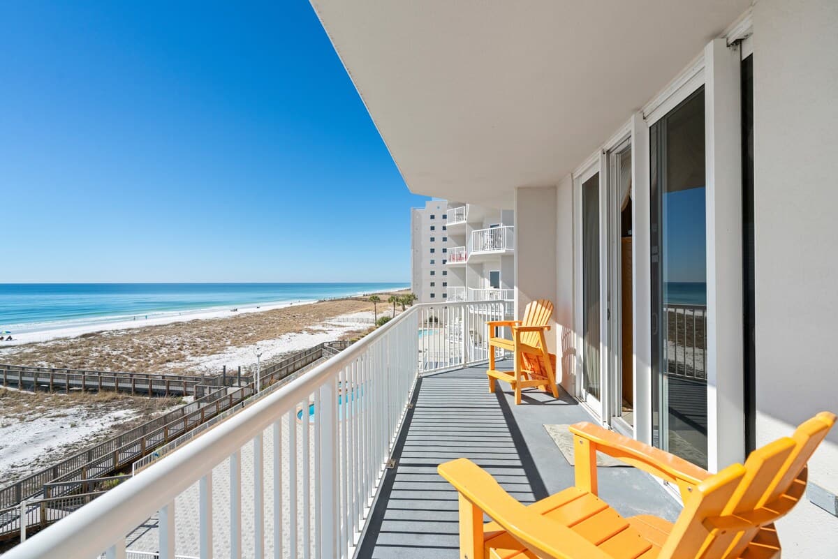 A spacious private balcony with gray flooring and white railings features two bright orange Adirondack chairs, offering panoramic views of the white sand beach, the ocean, and adjacent high-rise buildings.