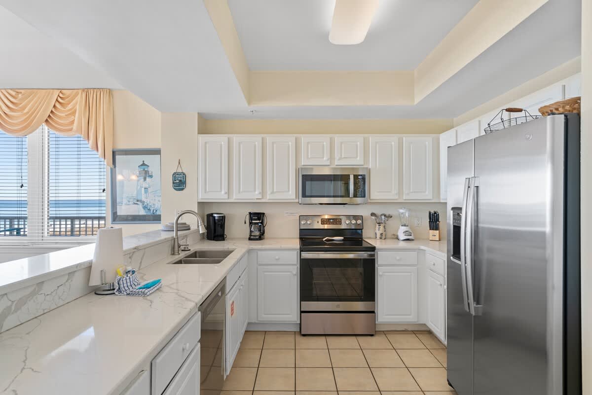 A modern kitchen with white shaker cabinets, stainless steel appliances including a microwave, oven, and refrigerator, and light marble-look countertops, all on light tile flooring.