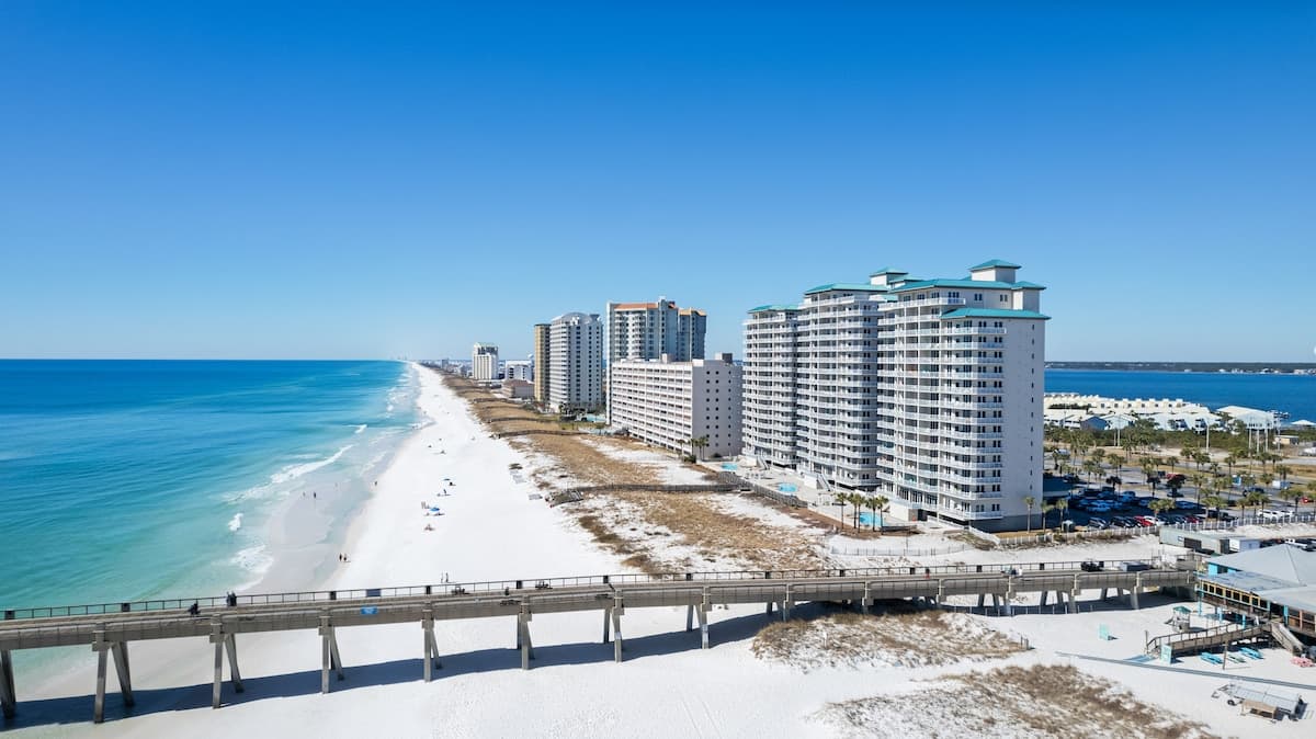 Aerial view along a pristine white sand beach, featuring several multi-story white condominium buildings with teal roofs, a long wooden pier, and the clear blue ocean stretching to the horizon.