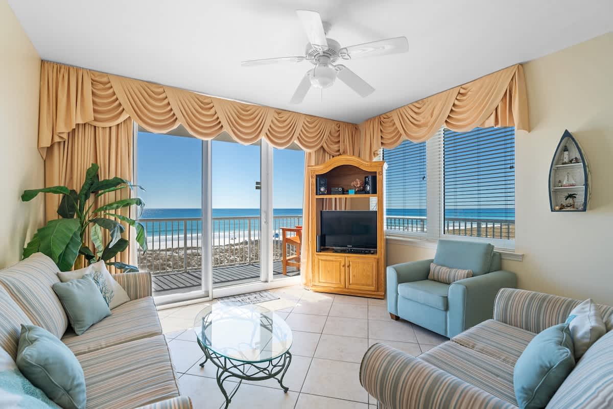 Bright living room with light-colored tile floors, two striped sofas, a glass coffee table, and a large sliding glass door leading to a balcony with panoramic ocean and pier views.