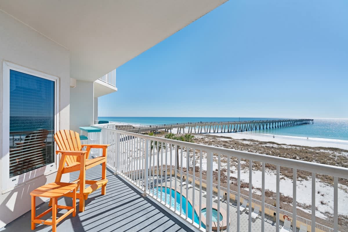 A private balcony features an orange Adirondack chair and a small blue bar table, overlooking a white sand beach, the ocean, a long pier, and a communal pool area below.