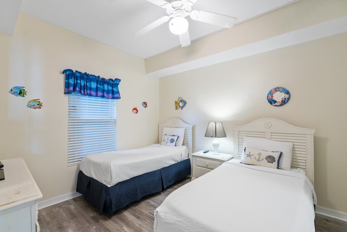 Cheerful bedroom with two white twin beds featuring blue bed skirts, light wood-look flooring, a ceiling fan, and playful fish-themed wall decor.