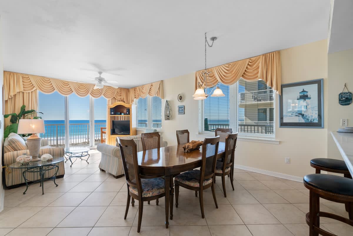 A bright living space with a striped sofa and armchair faces large windows offering panoramic ocean and pier views, with a dining area visible to the left and light tile flooring throughout.