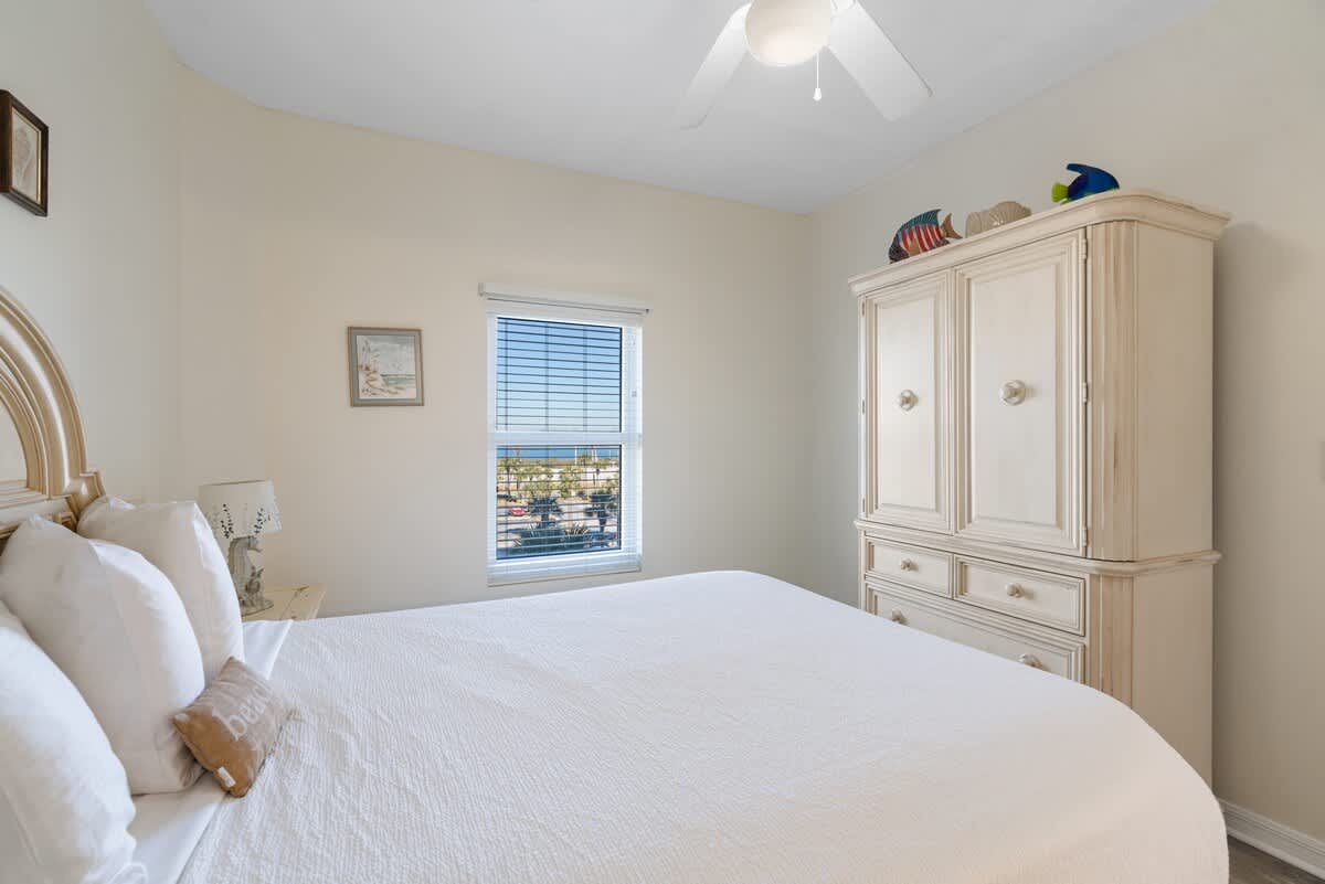 Another view of the queen bedroom, showcasing a large white armoire with drawers, a window with blinds, and subtle beach-themed decor.