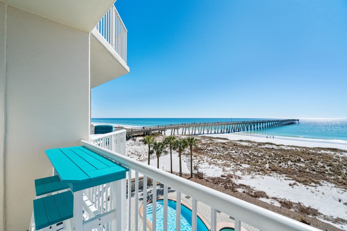 A vibrant blue bar table with matching stools is positioned on a private balcony, offering a clear view of the white sand beach, the ocean, a long pier, and a pool area with palm trees below.