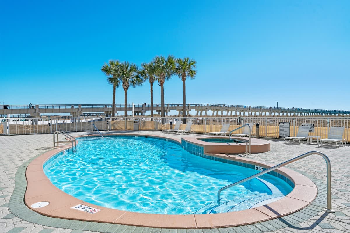 The communal kidney-shaped swimming pool and adjacent hot tub are set on a patterned paver deck, with several palm trees and a long pier visible in the background against a bright blue sky.