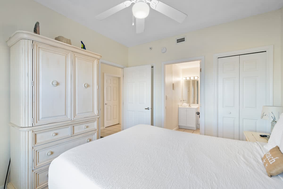 View of the queen bedroom with a white armoire and an open doorway leading to an en-suite bathroom, featuring light walls and a ceiling fan.