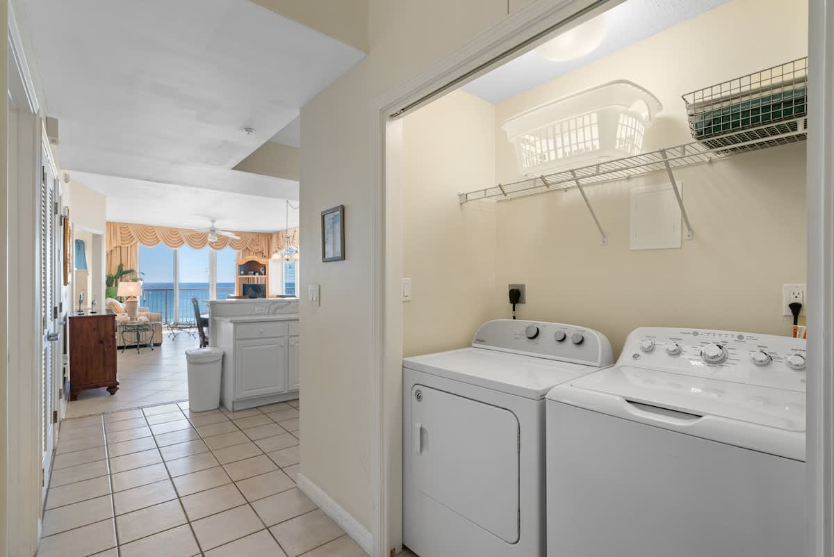 Functional laundry closet with a white top-loading washing machine and front-loading dryer, shelving above, and tiled flooring leading to an open living area with ocean views.