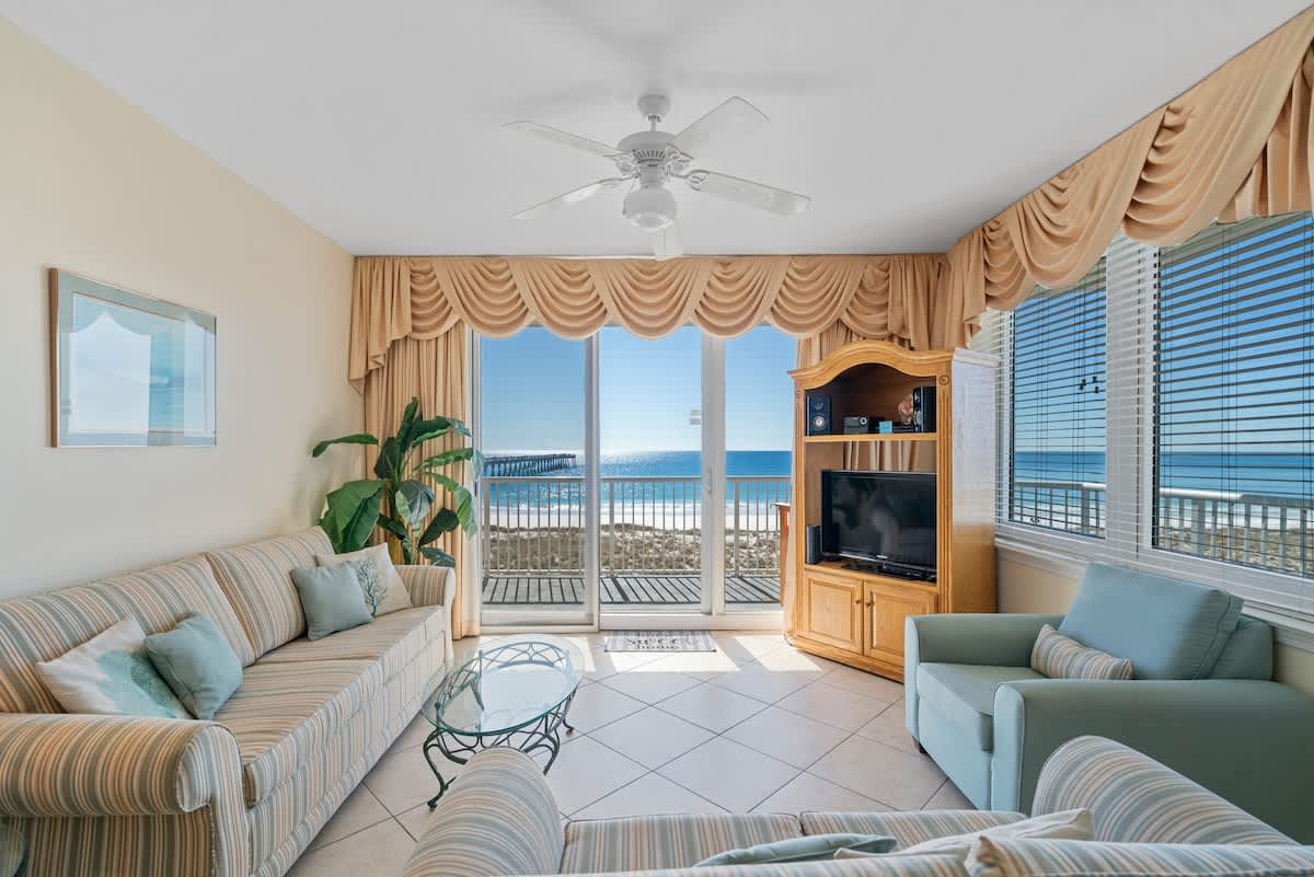 Spacious living room featuring light tile flooring, two comfortable striped sofas, a glass-top coffee table, and a large entertainment center, with a sliding glass door offering direct views of the ocean and pier.