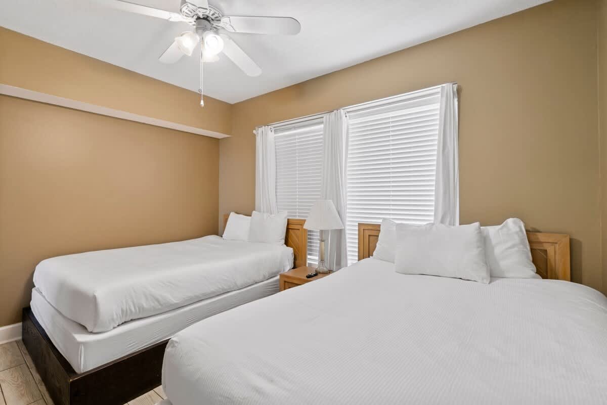 Another view of the bedroom featuring a queen bed and a twin bed, both dressed in crisp white linens, with light wood headboards and a shared nightstand. The room has warm beige walls, light tile flooring, and a ceiling fan.