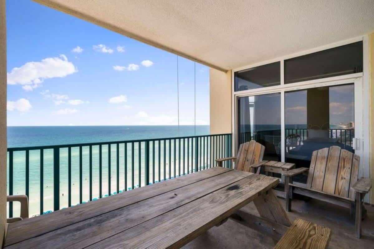 Spacious balcony featuring a rustic wooden picnic table and two Adirondack chairs, offering expansive, unobstructed views of the turquoise ocean and sandy beach below. A sliding glass door connects to an interior room with a visible bed.