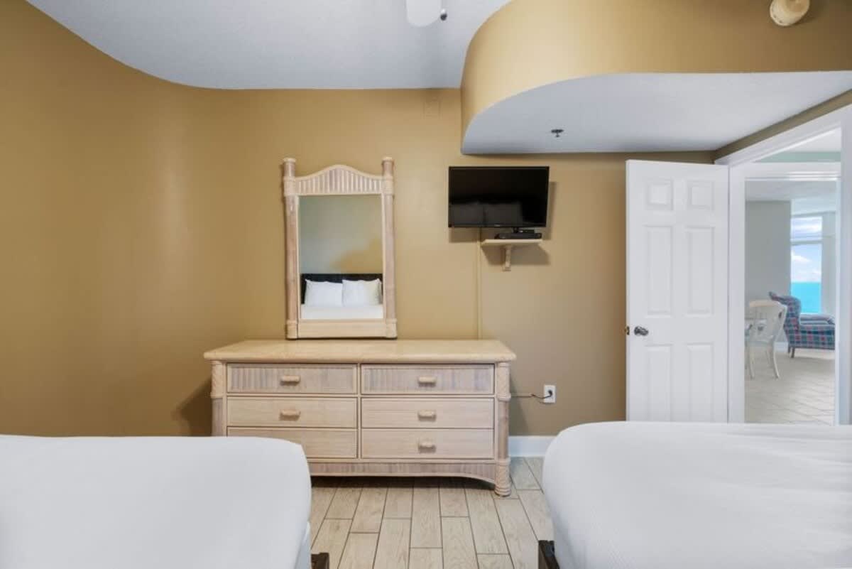 Second bedroom with two beds, light wood-look tile flooring, and a light-colored dresser with a mirror on top. A wall-mounted TV is positioned above the dresser, and a doorway leads to another area.