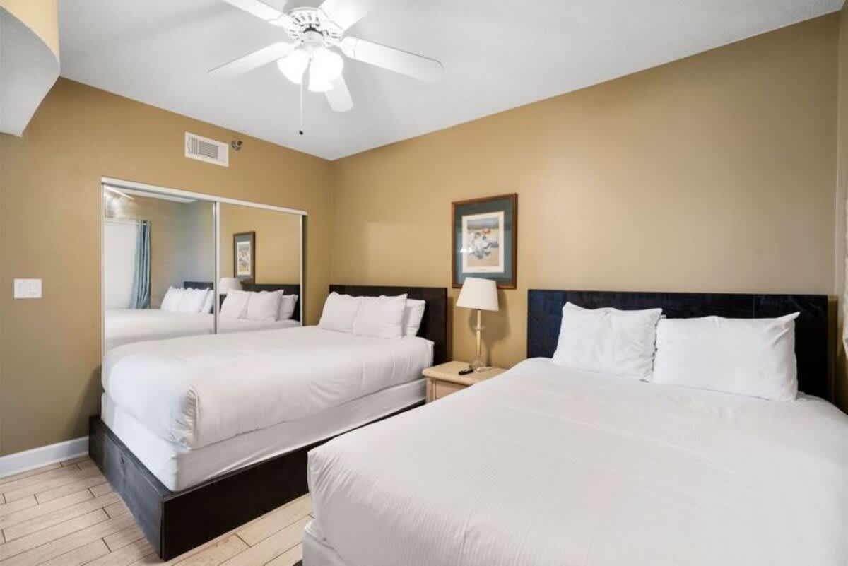 Second bedroom with two white-linened beds, light wood-look tile flooring, and a large mirrored closet door reflecting the room. A nightstand with a lamp is placed between the beds.