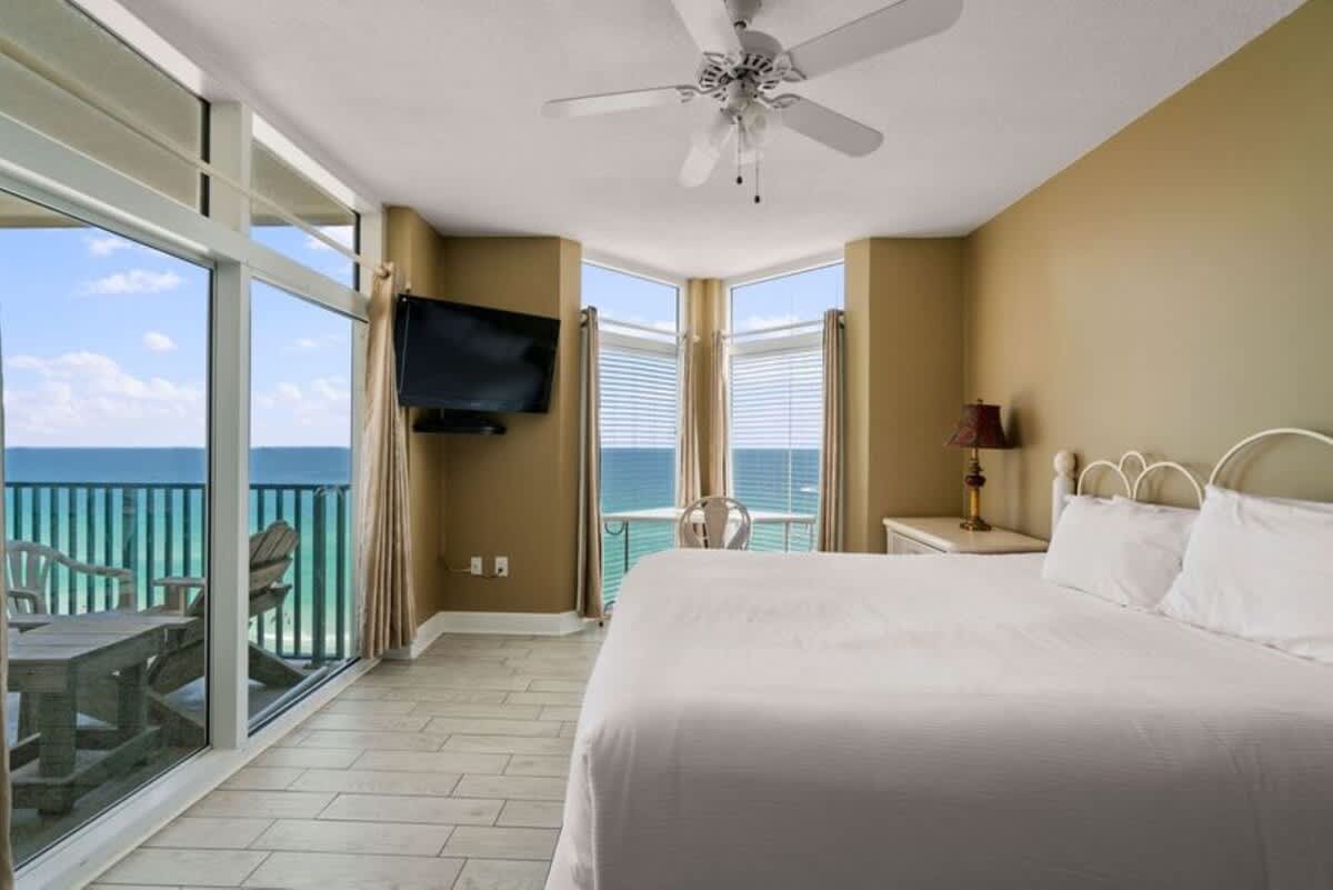 Master bedroom with a white bed, light wood-look tile flooring, and tan walls, featuring a large sliding glass door leading to a balcony with outdoor seating and expansive ocean views. A wall-mounted TV is visible on the left.