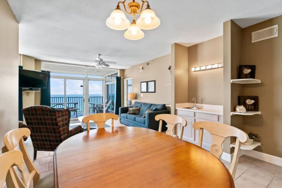 Dining area with a light wood oval table and four chairs, featuring a small wet bar with shelving on the adjacent wall. The room opens to a living area with a blue sofa and large windows offering ocean views.