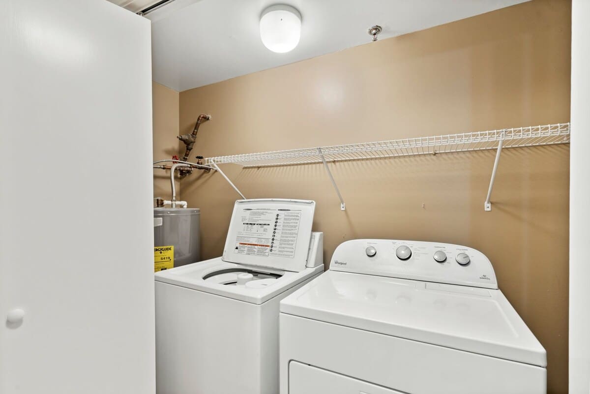 Compact laundry area with a white top-loading washing machine and a white front-loading dryer, situated beneath a wire shelf for storage. A water heater is visible in the corner, and the walls are painted a warm beige.