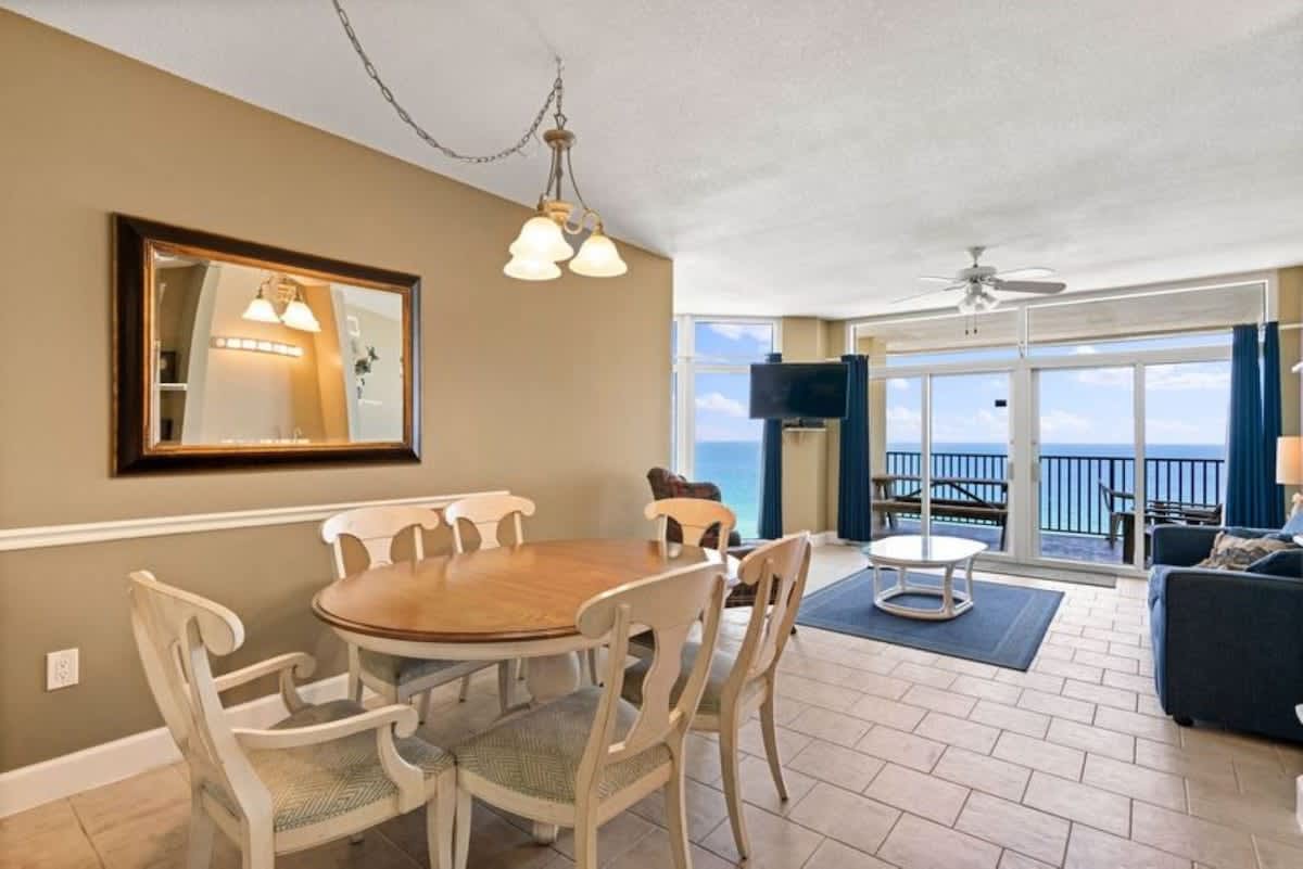 Elegant dining area with a light wood oval table and six white chairs, illuminated by a three-light chandelier. A large framed mirror hangs on the wall, and the space opens to a living room with ocean views in the background.