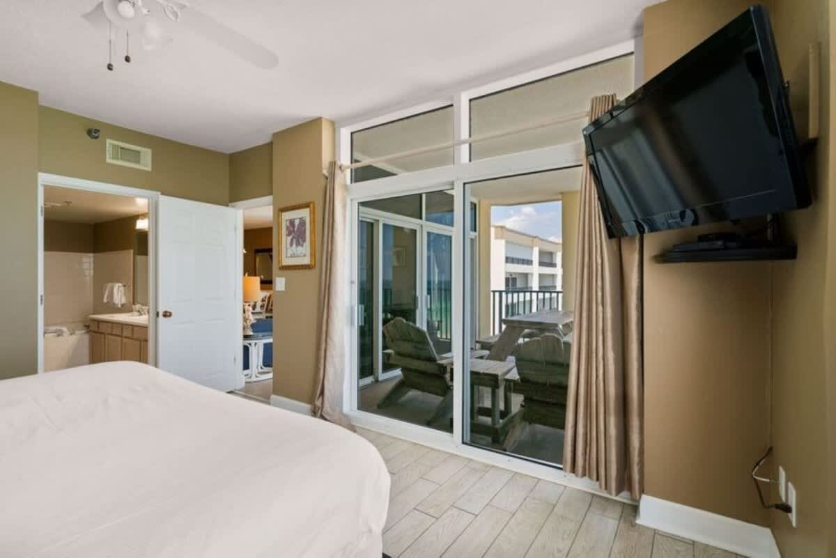 Master bedroom with a white bed and light wood-look tile flooring, featuring a large sliding glass door to a balcony with ocean views, and an open doorway leading to an en-suite bathroom. A wall-mounted TV is visible on the right.