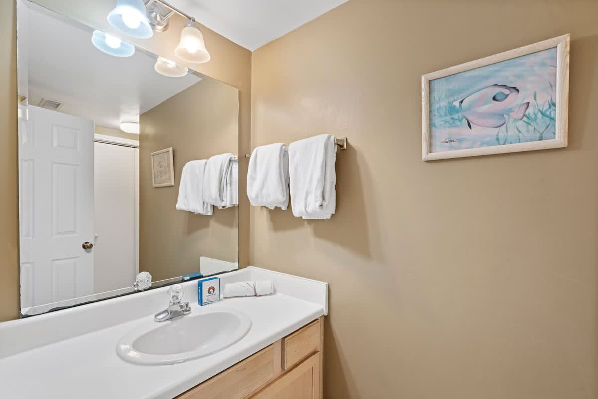 Bathroom vanity with a white countertop, integrated sink, and light wood cabinetry, illuminated by a multi-bulb light fixture above a large mirror. White towels are neatly folded on a rack, and a framed fish artwork adorns the warm beige wall.