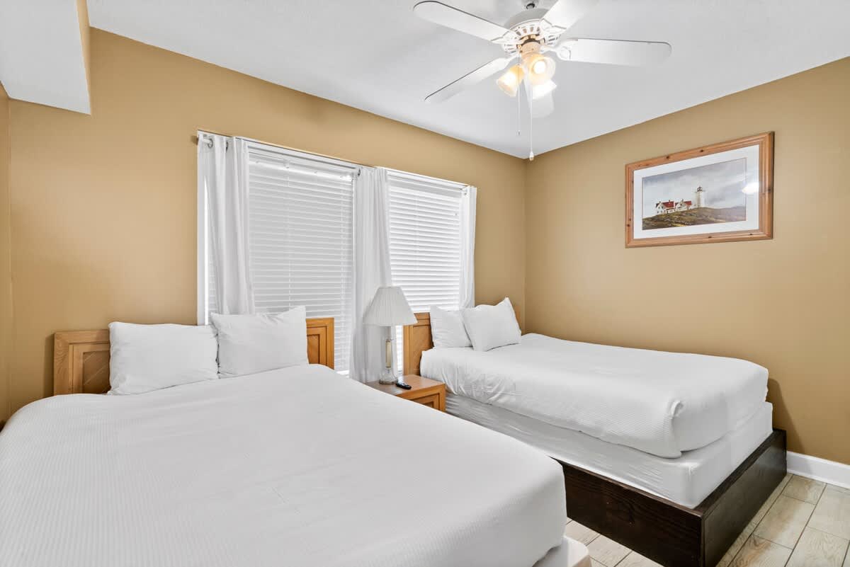 Bright bedroom with two beds, one queen and one twin, both with white bedding and light wood headboards, set against warm beige walls. A ceiling fan with lights hangs overhead, and large windows with white blinds and curtains provide natural light.
