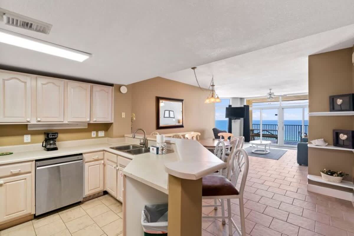 Open-concept kitchen with white cabinets and light countertops, including a breakfast bar with three stools. The kitchen overlooks the dining and living areas, which feature tiled floors and large windows with ocean views.