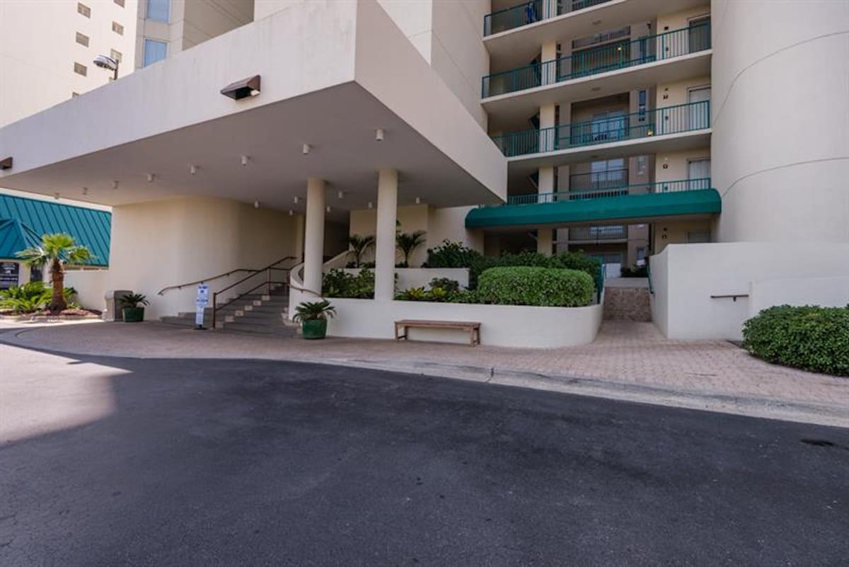 The grand entrance to the condominium building features a covered drive-through portico with multiple support columns, leading to a set of stairs and a ramp. Lush green landscaping, a wooden bench, and potted plants adorn the entrance area.