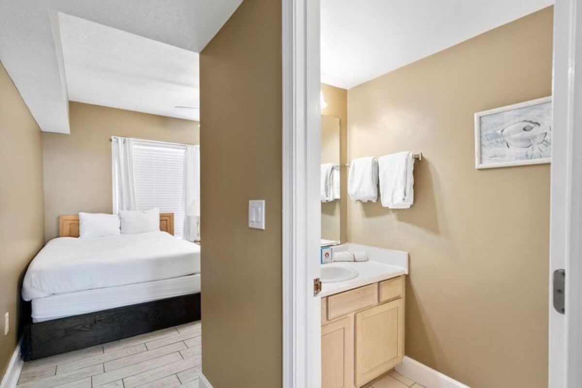 View from a bathroom vanity area into a bedroom, featuring a bed with white bedding and a light wood headboard. The foreground shows a bathroom vanity with a white sink and mirror, all set against warm beige walls and light tile flooring.