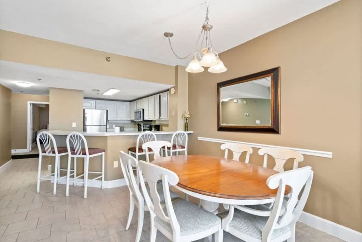 Dining area featuring a light wood oval table with six white chairs, positioned under a three-light chandelier. The space connects to a kitchen with a breakfast bar and white cabinetry, all set on light tiled flooring.