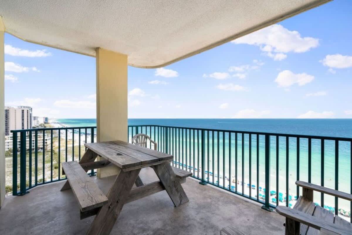 Wide-angle view of a large balcony furnished with a weathered wooden picnic table and chairs, providing panoramic vistas of the vibrant blue ocean and distant coastline. A sturdy dark metal railing secures the perimeter.
