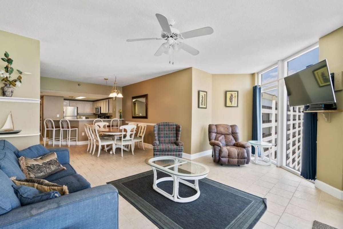 Open-concept living area with tiled floors, featuring a blue sofa, a plaid armchair, and a brown recliner. The space flows into a dining area and kitchen, all bathed in natural light from large windows offering partial ocean views.