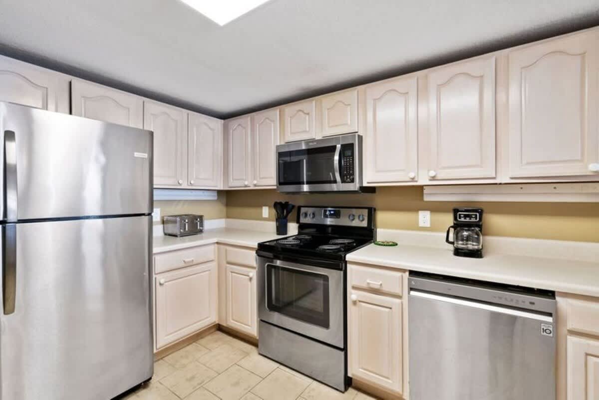 Close-up view of a well-equipped kitchen featuring white upper and lower cabinets, light countertops, and stainless steel appliances including a refrigerator, microwave, oven, and dishwasher. A coffee maker and toaster are also visible on the counter.