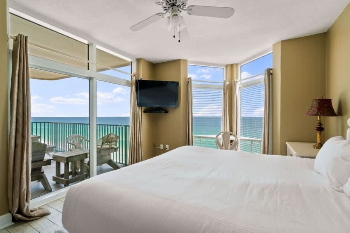 Master bedroom with a white bed, light wood-look tile flooring, and tan walls, offering direct access to a balcony with wooden outdoor furniture and stunning ocean views through a large sliding glass door. A wall-mounted TV is on the left.