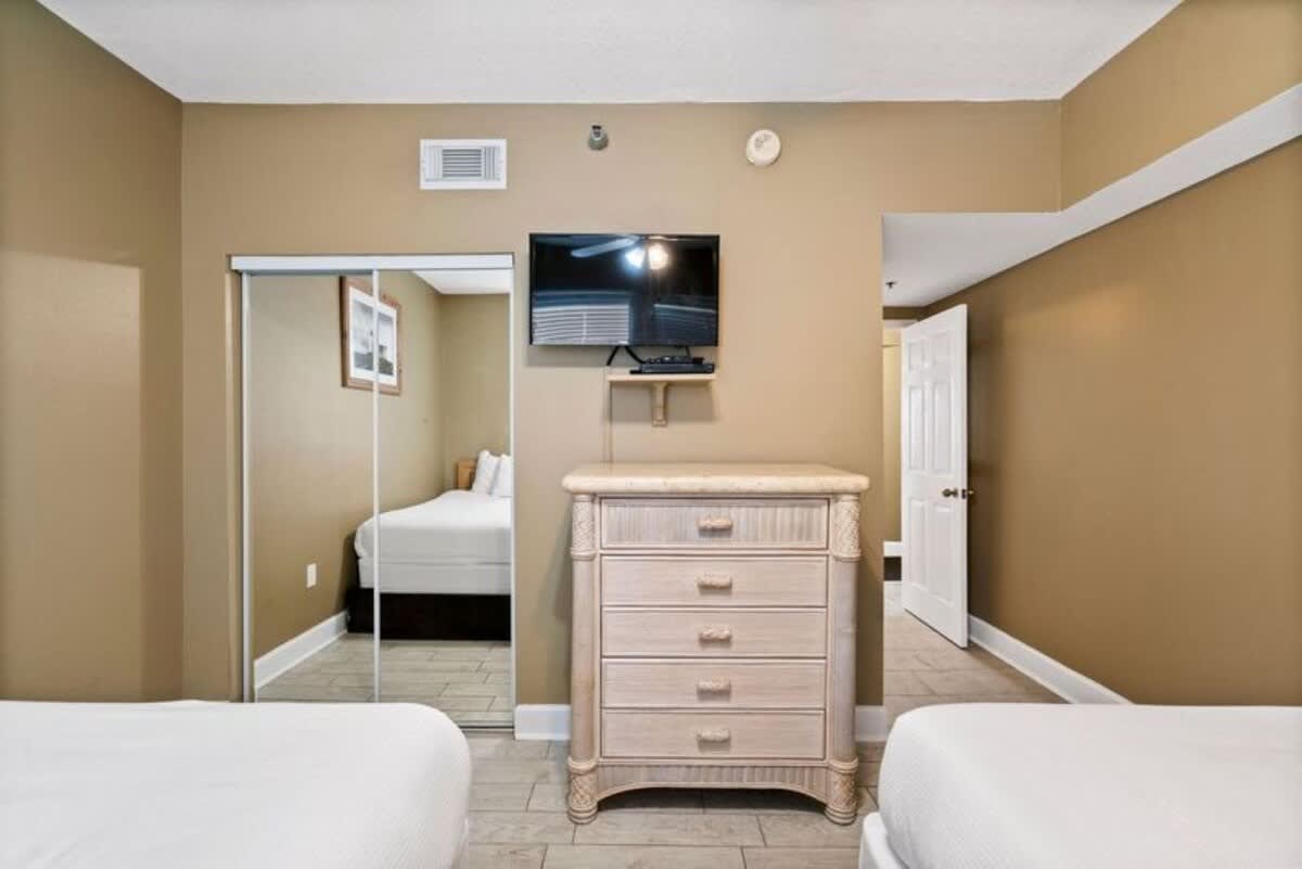 Bedroom view showcasing a light-colored wicker dresser with five drawers and a wall-mounted flat-screen TV above it. Mirrored closet doors reflect the two beds in the room, which has warm beige walls and light tile flooring.