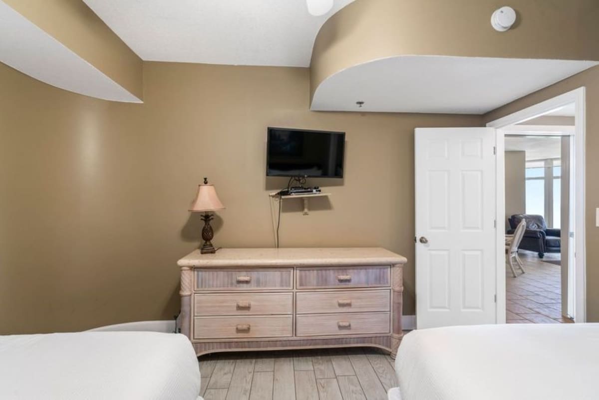 Bedroom view showcasing a light-colored dresser with multiple drawers, a wall-mounted television, and an open doorway leading to another room, with two beds partially visible.