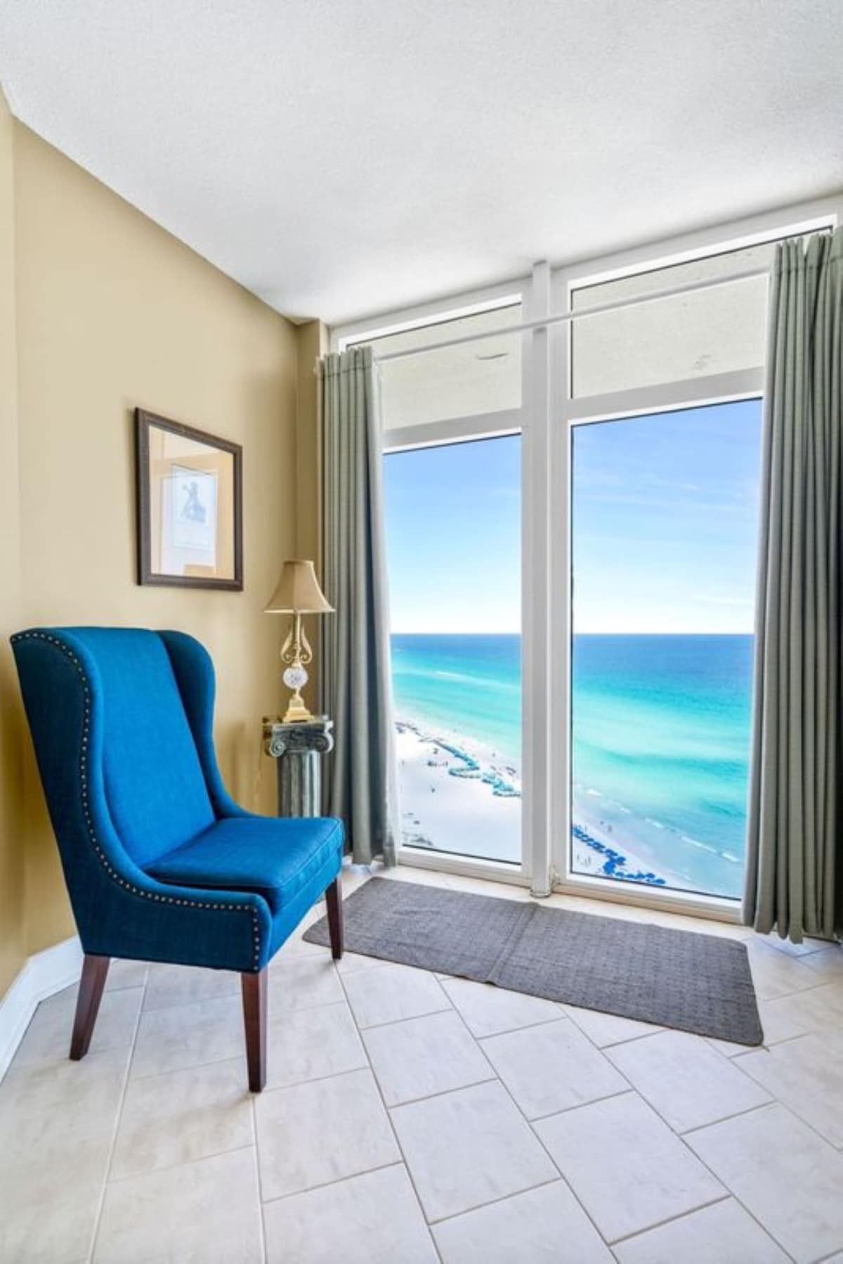 Cozy sitting nook with light tan walls and tile flooring, featuring a vibrant blue upholstered armchair with nailhead trim. Large windows offer a breathtaking view of the white sand beach and turquoise ocean.