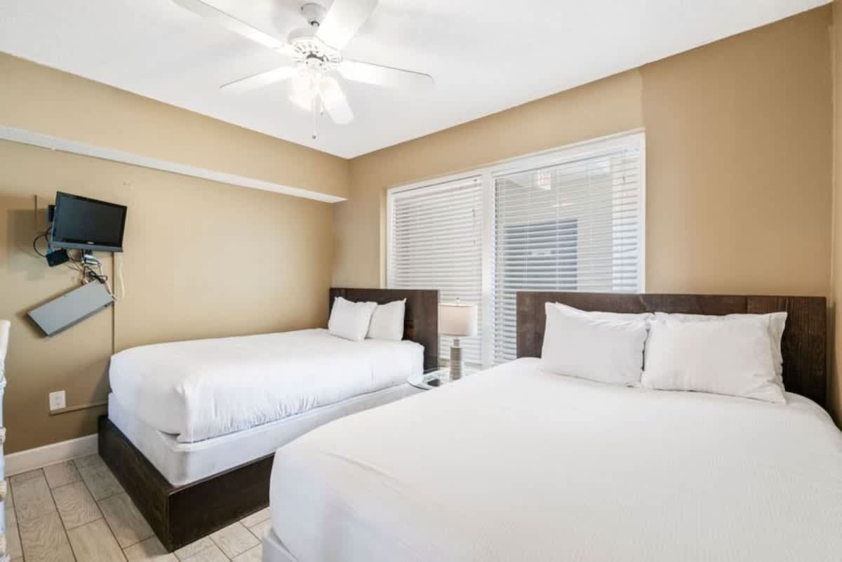 Bedroom featuring a queen bed and a twin bed with dark wood frames and white linens, light wood-look tile flooring, and a large window with white blinds.