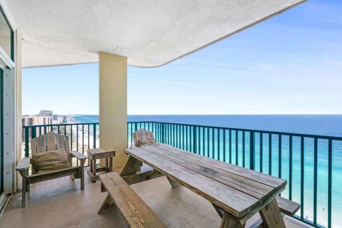 Expansive balcony featuring a large wooden picnic table and two wooden Adirondack-style chairs, all set against a stunning backdrop of the ocean and coastline.
