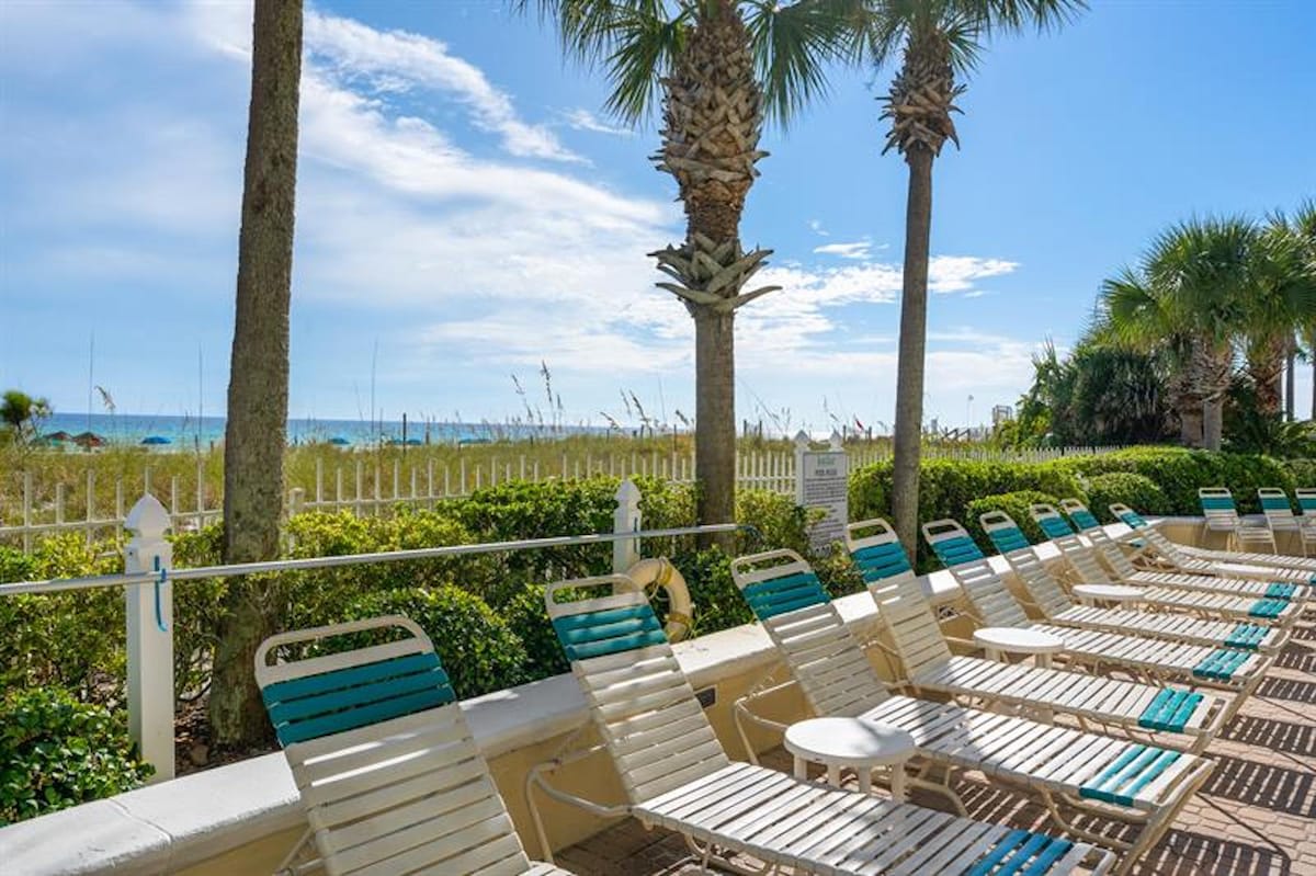 A row of white and teal lounge chairs is neatly arranged on a paved area, offering direct views of the white sand beach and the sparkling ocean, framed by lush greenery and palm trees.