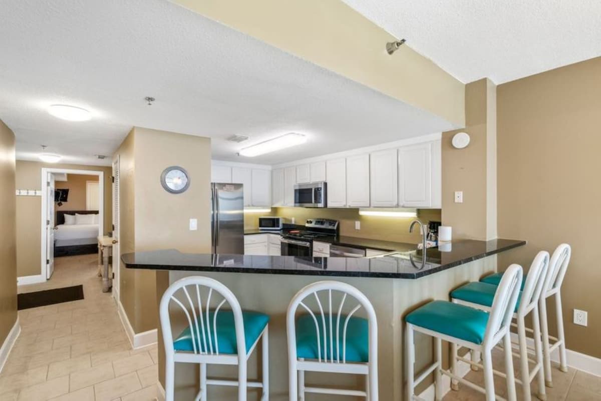 Spacious kitchen featuring a dark granite breakfast bar with four teal-cushioned white bar stools, and a view of the stainless steel appliances and white cabinets.