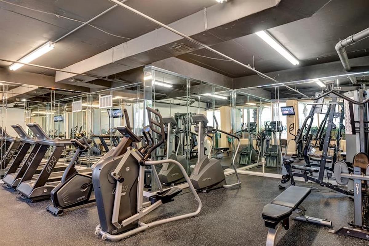 A well-equipped fitness center features multiple treadmills, elliptical machines, and weightlifting equipment, with mirrored walls reflecting the exercise area under fluorescent lighting.