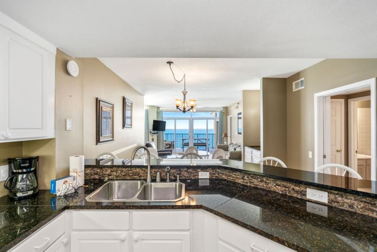 Kitchen counter with a double stainless steel sink, coffee maker, and paper towel holder, offering an open view over the breakfast bar to the dining and living areas with ocean views.