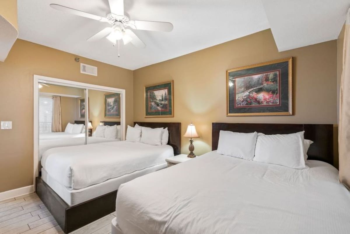 View of a bedroom with two beds, one queen and one twin, both with dark wood frames and white bedding, alongside a mirrored closet and light wood-look tile flooring.