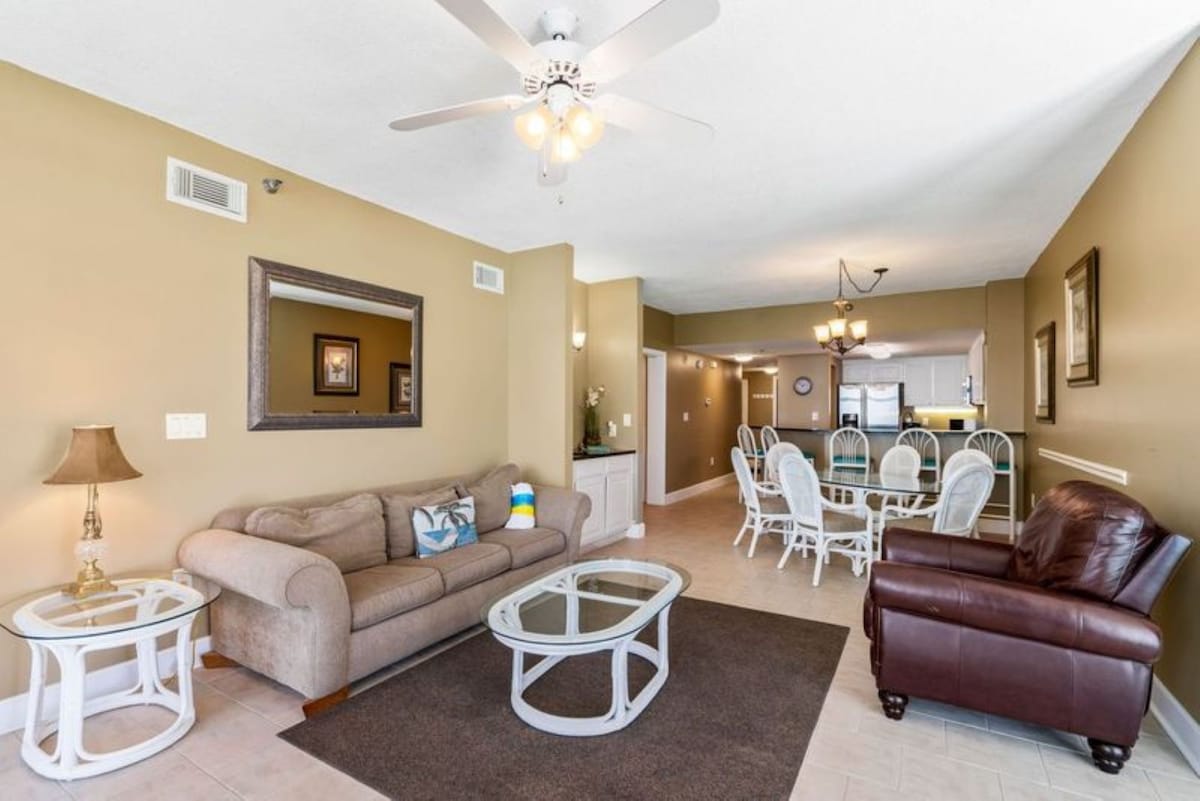 Spacious open-concept living area featuring a plush brown sofa, a leather armchair, and a glass coffee table on a dark area rug, with a ceiling fan and a large mirror, flowing into the dining space.