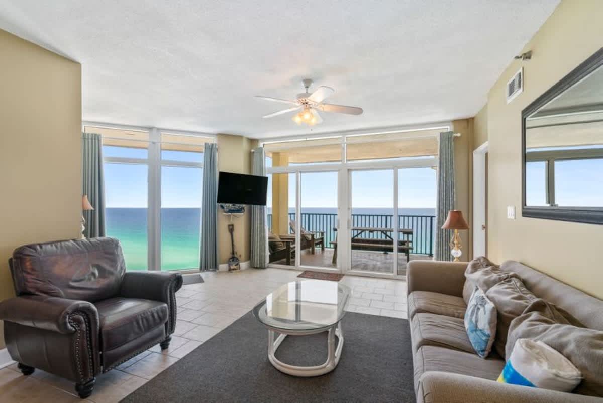 Spacious living room with light tan walls, tile flooring, and floor-to-ceiling windows offering panoramic ocean views. Features a dark leather armchair, a light-colored sofa, and a glass coffee table on a dark rug.