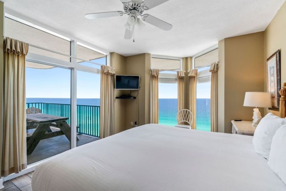 Master bedroom with a king bed, wood-look flooring, and direct access to a balcony through sliding glass doors. The balcony features a wooden picnic table, offering clear views of the turquoise ocean.