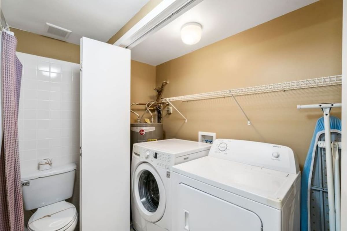 A compact laundry area features a white front-loading washing machine and dryer, a water heater, and a toilet tucked behind a partial wall, all set against light brown walls.
