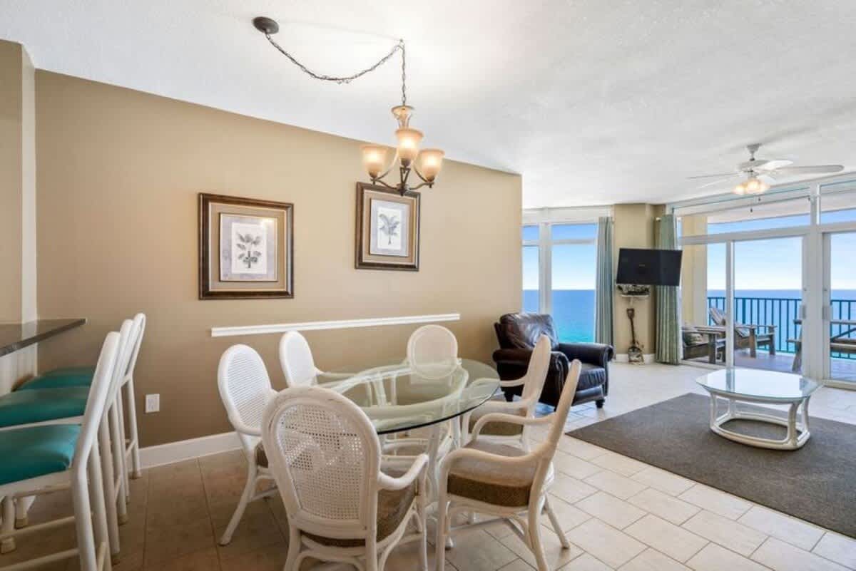 Elegant dining area featuring a round glass-top table with white wicker chairs, illuminated by a classic chandelier, with an open layout leading to the living room and ocean-facing balcony.