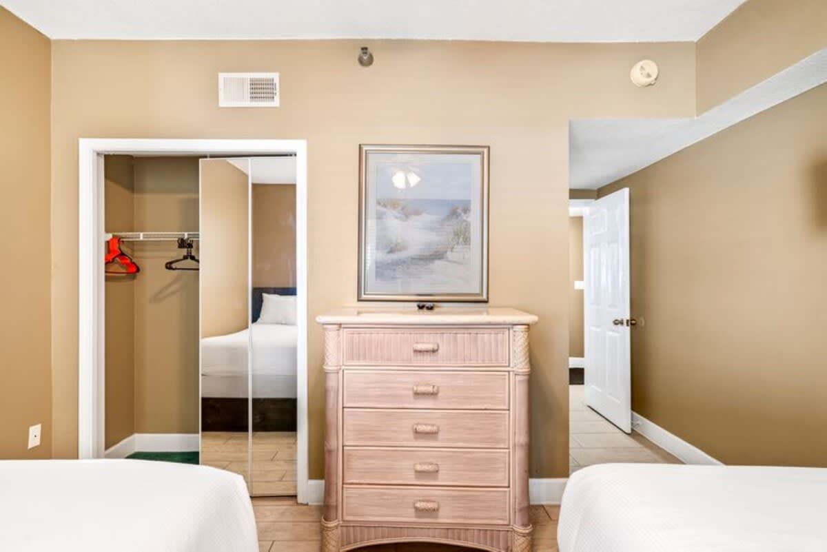 Bedroom view with a light wood dresser and a framed artwork above it, a mirrored closet, and two beds with white linens partially visible on either side.