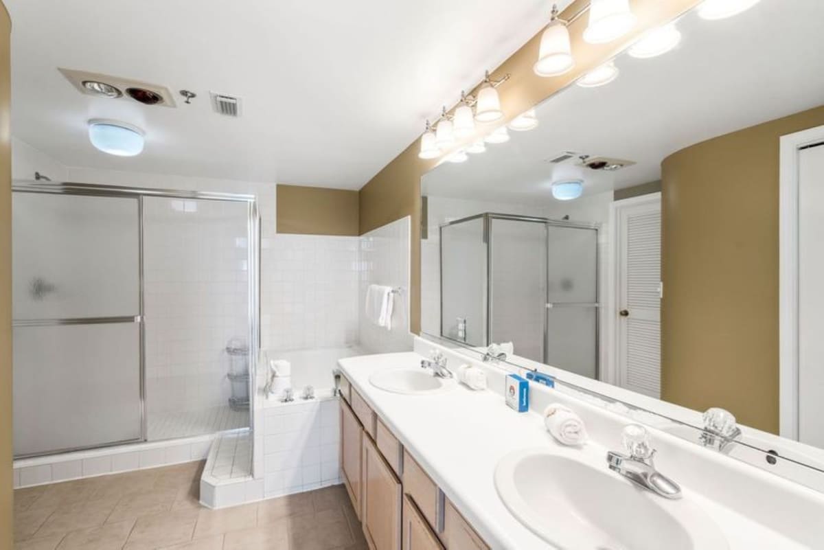 Spacious bathroom featuring a double vanity with white countertops and light wood cabinets, a large mirror with vanity lighting, and a separate shower with sliding glass doors next to a built-in bathtub.